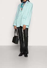 Blazer oversize vert menthe avec deux poches frontales, porté sur un haut à motifs. Pantalon en similicuir noir et petit sac à main noir.