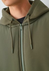 Olivově zelená mikina s hladkou texturou, vybavená zipem a nastavitelnou šňůrkou. Kapuce je jemně podšitá, což zvyšuje pohodlí.