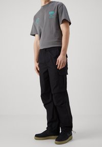 Man in een grijs graphic t-shirt, zwarte cargobroeken met zijzakken, en zwarte sneakers met beige zolen, staand tegen een effen achtergrond.