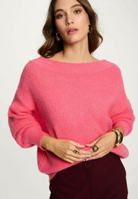 Maglione rosa oversize a maglia con scollo a barca, texture a coste e maniche ampie. Abbinato a pantaloni scuri e accessori dorati.