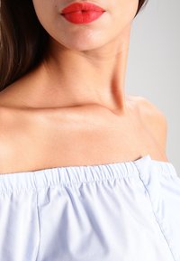 Světle modrý top s odhalenými rameny a elastickým shrnovacím výstřihem; hladká textura a lehká látka. Výrazné klíční kosti a detail červené rtěnky.