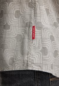 Close-up van de zoom van een grijs patroondem shirt met het rode label "JOOP! JEANS" boven de naad van donkerdenim jeans.