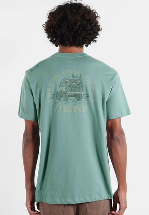 Personne aux cheveux bouclés portant un t-shirt vert montrant un graphique arrière d'un véhicule transportant un vélo et le texte "Having Good Times Oxbow."