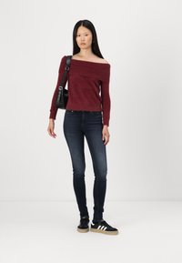 Bordo pulover bez ramena od mekog materijala, uparen s tamnim uskim trapericama i crnim tenisicama s bijelim naglascima. Crna torba preko ramena.
