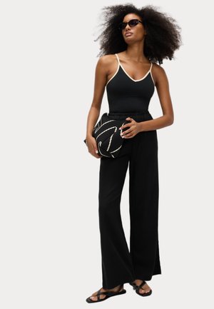 Zwarte jumpsuit met lichte afwerking, brede zwarte broek en gestrikte zwarte sandalen. Houdt een patroon zwart clutch met witte accenten vast.