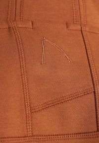 Gros plan sur un tissu couleur rouille avec des coutures détaillées et un petit logo brodé subtil en fil assorti.