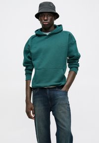Zielona bluza z kapturem i kieszenią z przodu, w połączeniu z ciemnoniebieskimi dżinsami. Czarny kapelusz typu bucket dodaje kontrastu. Gładki materiał z delikatnym przeszyciem.
