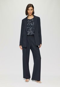 Traje de marinero ajustado con una chaqueta estructurada y pantalones de pierna ancha, combinado con una blusa negra que presenta un patrón de hojas blancas y tirantes finos.
