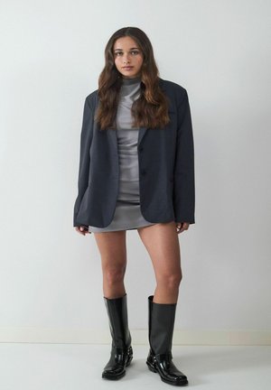 Vestido mini de satén gris, con una chaqueta entallada oscura encima. Botas negras de charol hasta la rodilla. Texturas suaves, diseño estructurado y colores neutros.