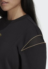 Juodas džemperis su apvalia iškirpte, raukšlėmis apdirbtais rankogaliais, tekstūruotu juodu tinklinio audinio akcentu ant pečių ir auksinio tono logotipu.