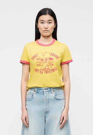 Tânără cu păr negru drept poartă un tricou galben cu margini roz și un desen cu un șoarece de desene animate, asortat cu blugi albaștri deschis, cu talie înaltă.