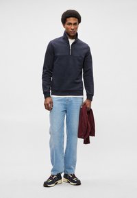 Felpa mezza zip, in misto cotone, jeans azzurri, sneakers sportive bianco e nero. Tiene una felpa bordeaux.