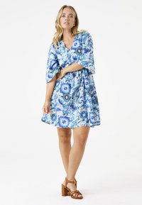 Kleid mit kurzen Ärmeln in einem blauen Blumenmuster, mit V-Ausschnitt, geraffter Taille und fließender Silhouette. Getragen mit braunen High-Heel-Sandalen.