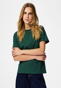 Jonge vrouw met lang blond haar, gekleed in een groen shirt met korte mouwen en een donkere blauwe spijkerbroek, met gekruiste armen tegen een effen witte achtergrond.