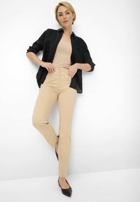 Pantalons beiges taille haute au design ajusté, associés à un débardeur beige clair et une chemise noire à boutons ample. Flats noirs à bout pointu.