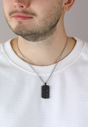 Schwarze Anhänger-Halskette mit filigranen Details, an einer silbernen Kette getragen, über einem strukturierten weißen Pullover.
