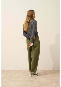 Szary sweter, luźne oliwkowe spodnie i szaro-białe sneakersy. Długie włosy, ręce złożone za plecami, stojący przy neutralnej ścianie.