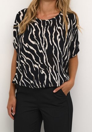 Vrouw draagt een kort mouwen blouse met zwart-wit zebraprint, gecombineerd met een zwarte broek, met één hand in de zak.