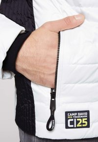 Giacca isolante bianca e nera con pannello laterale testurizzato, tasca con zip e etichetta Camp David. Presenta una finitura liscia e trapuntata.