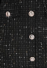 Chaqueta negra texturizada con acentos en hilo dorado y blanco, con dos filas horizontales de botones plateados brillantes en la parte delantera.