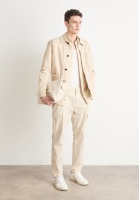 Chaqueta de lona beige con cierre de botón, combinada con pantalones cargo a juego y zapatillas blancas. Modelo sosteniendo un clutch beige. Diseño minimalista.