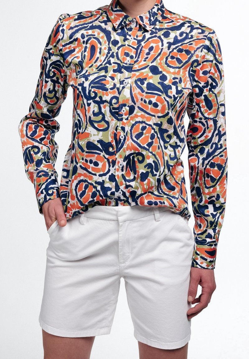 Personne portant une chemise à motifs abstraits colorés avec des formes bleu marine, orange et olive, associée à un short blanc.