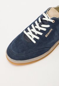 Marineblå semsket sneakers med hvite snørebånd, perforert tå og beige detaljer. Har en såle av gummi og metallisk logo på siden.