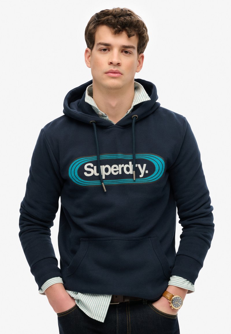Granatowy kapturowy sweter z dużym logo "Superdry", kangurzą kieszenią i sznurkami do regulacji. Noszony na jasnej, wąsowanej koszuli.