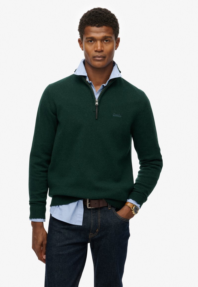 Grøn strikket sweater med lynlås krave over en lyseblå skjorte. Mørke jeans og et brunt bælte med et ur fuldender outfittet.