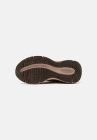 Semelle d'une basket marron et beige montrant un motif de grip texturé avec des rainures de flexion et le nom de la marque embossé près de la voûte plantaire.
