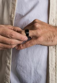 Schwarzer Bandring an einem Finger, mit flachem oberen Teil und glatter Oberfläche. Die Hände sind positioniert, eine passt den Ring an. Texturiertes Hemd sichtbar.