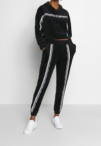 Czarny dres z weluru, składający się z krótkiej bluzy z kapturem oraz spodni z elastycznym pasem. Posiada białe boczne paski z brandingiem. Połączony z białymi sneakerami.