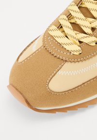 Brune semsket sneaker med gule mønstrede snørebånd, glatte beige stoffpaneler, hvite sting og såle av teksturert brun gummi.