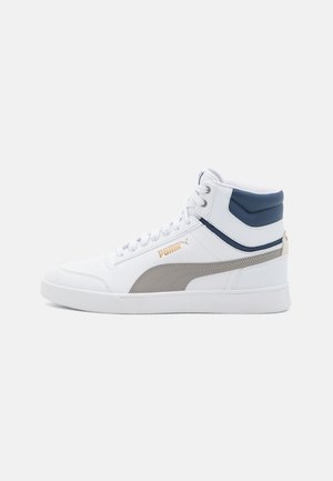 Zapatilla blanca de caña alta Puma con franja gris, detalles en azul marino en el cuello y logo dorado en el lateral, mostrada en perfil izquierdo sobre un fondo liso.