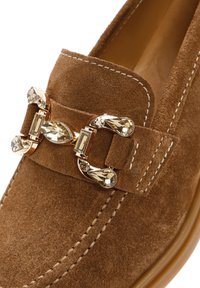 Mocassin en daim marron avec boucle dorée décorative ornée de pierres précieuses transparentes et facettées et coutures blanches contrastées.