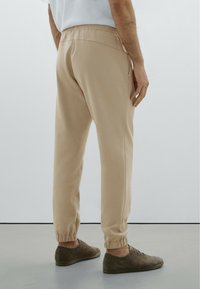 Beige joggerbroek van een textuurstof, met een elastische tailleband en geribbelde boorden. G gedragen met bruine schoenen, wat een ontspannen fit van achteren laat zien.