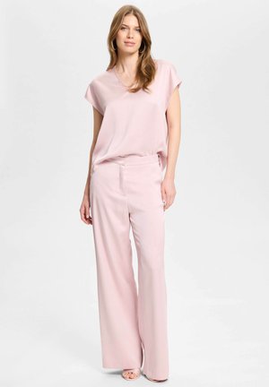 Jonge vrouw met lang haar met grote oorbellen, een lichtroze korte mouwen blouse, broek met wijde pijpen en open sandalen met hak, staand.