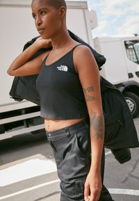 Person with shaved head wearing a black cropped tank top and pants, holding a black jacket over shoulder, standing near a parked white truck.