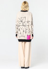 Pull crème avec des motifs de chats dessinés en noir et rose ; coupe ample avec poignets et ourlet noirs ; texture douce ; associé à un pantalon large beige clair.
