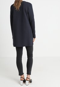 Granatowy, teksturowany, długi płaszcz z długimi rękawami, w połączeniu z czarnymi obcisłymi dżinsami i srebrnymi butami na słupku. Widok z tyłu pokazuje długość i dopasowanie płaszcza.