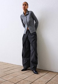 Žena v šedej zapínacej cardigan, širokých pruhovaných nohaviciach a čiernych špicatých topánkach, stojí proti bielej textúrovanej stene a dlažbe.