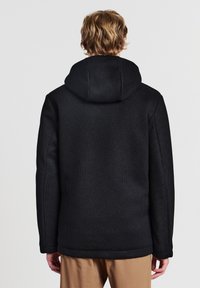 Persona che indossa una giacca a cappuccio nera con texture e pantaloni beige, vista da dietro su uno sfondo chiaro e uniforme.