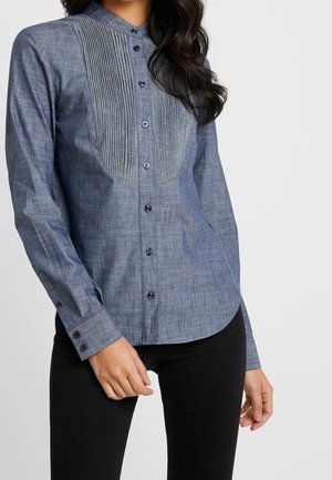 Woman wearing a long-sleeve blue button-up shirt with vertical pleats on the chest, paired with black pants.