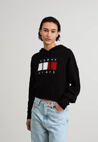 Crna hoodie s pletenim uzorkom i skraćenim dizajnom, koja ima šareni logo u crvenoj, bijeloj i mornarsko plavoj boji. Nosena s svijetloplavim trapericama.