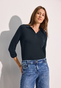 Musta pitkähihainen poolopaita V-kauluksella ja käännetyillä hihoilla, yhdistettynä vaaleansinisiin kulutetun näköisiin suorin lahkein farkkuihin, joissa on erottuva ommel.