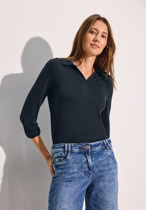 Schwarzes Langarm-Polohemd mit V-Ausschnitt und umgeschlagenen Manschetten, kombiniert mit hellblauen, verwaschenen, geraden Jeans mit kontrastierenden Nähten.