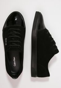 Černé semišové nízké tenisky s lesklou špičkou, černými tkaničkami a gumovou podešví, zobrazené z horního a bočního pohledu na bílém pozadí.