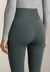 Magas derekú, bordázott, sötétzöld leggings, rugalmas anyagból készült, sima textúrával és diszkrét márkajelzéssel a derékrész felett.