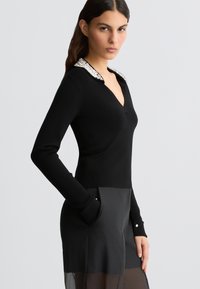 Maglione nero a maniche lunghe con scollatura a V e colletto di perline. Texture liscia, design aderente, abbinato a pantaloni in tessuto trasparente neri.