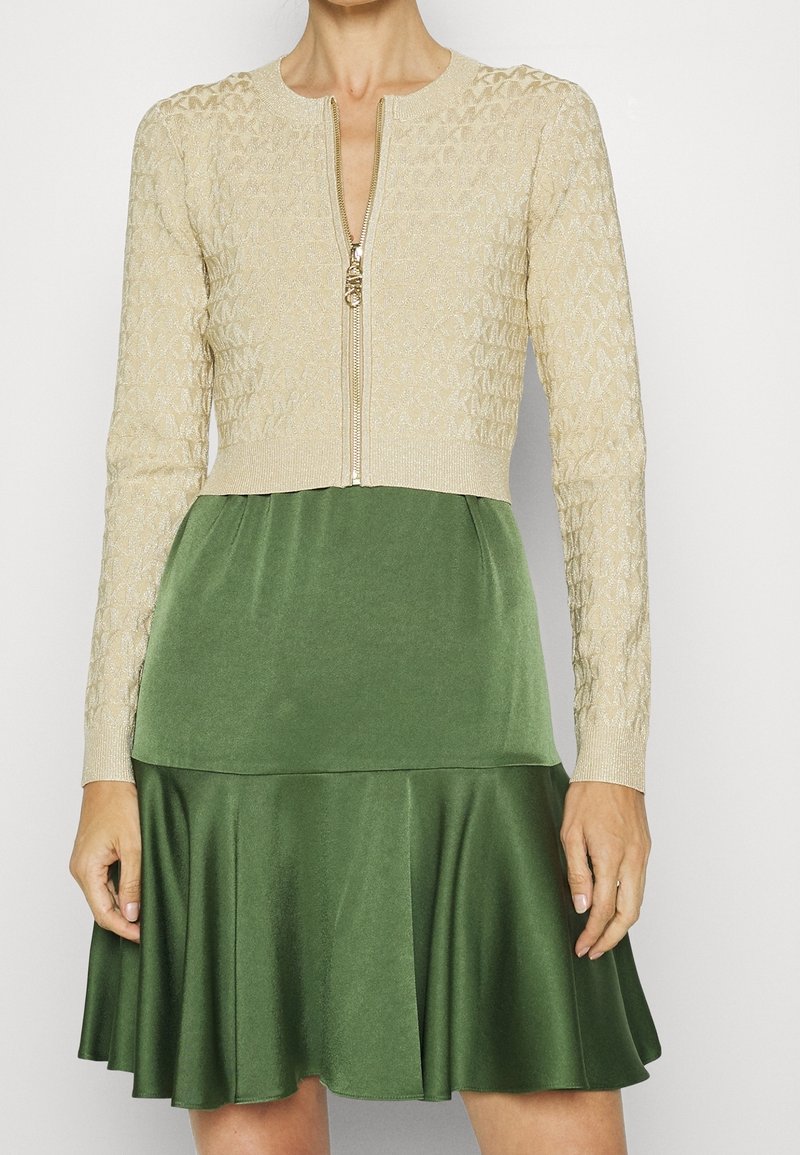 Cardigan lavorato a maglia beige con motivo testurizzato, chiusura con zip; abbinato a un vestito verde svasato in tessuto liscio, modello a ginocchio.